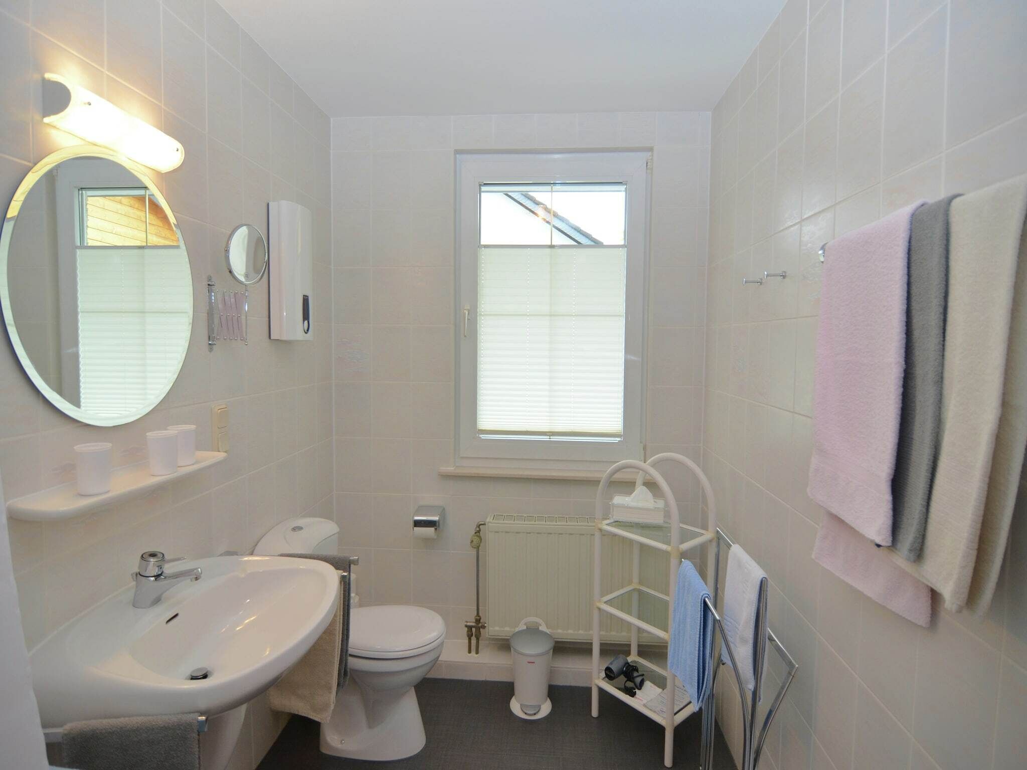 Une petite salle de bain propre, équipée de toilettes, d’un lavabo et d’un miroir. Une fenêtre laisse entrer la lumière naturelle, et un porte-serviettes contient plusieurs serviettes.