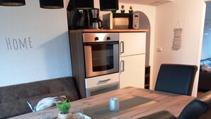 Moderni keittiö, jossa on sisäänrakennettu uuni ja mikroaaltouuni, ruokapöytä tuoleineen sekä seinälle kiinnitetty hylly, jossa on keittiötarvikkeita.