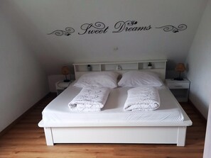 Een net opgemaakt bed met witte lakens en twee opgerolde handdoeken, een wit hoofdbord, twee nachtkastjes met lampen, en een wanddecoratie met de tekst 'Sweet Dreams'.