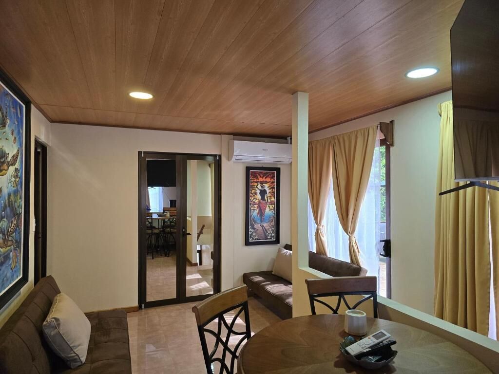 A spacious living room with a wooden ceiling, a dining area with a table and chairs, a sofa, and a large painting on the wall.