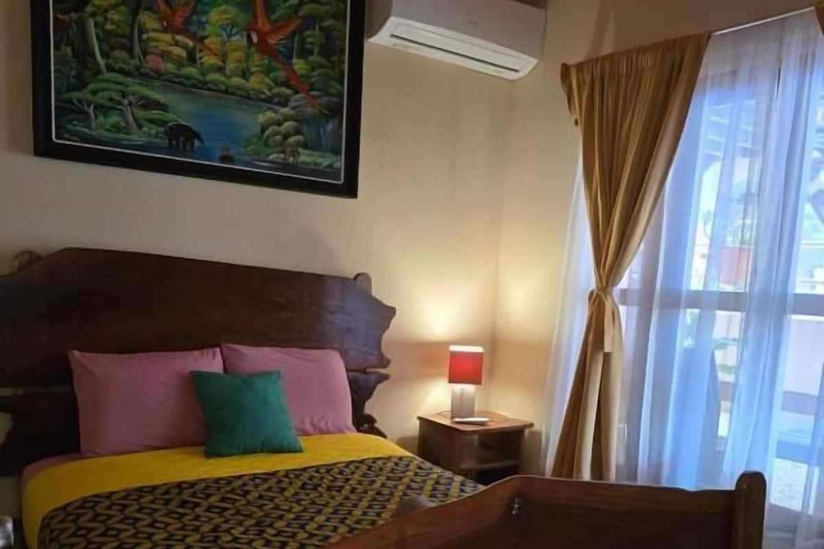 Une chambre avec un lit en bois, des coussins colorés, un tableau représentant une scène de jungle, un climatiseur, une lampe de chevet et une fenêtre avec des rideaux.