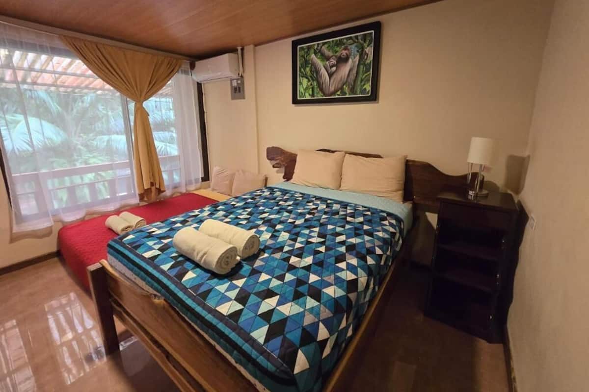 Une chambre à coucher avec un lit, une table de chevet, une fenêtre avec des rideaux et un tableau accroché au mur.
