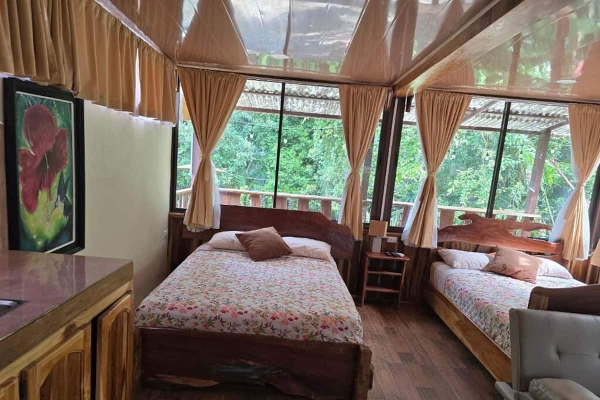 Une pièce avec deux lits en bois, une petite table et une chaise. Il y a une fenêtre avec des rideaux et un tableau encadré au mur.