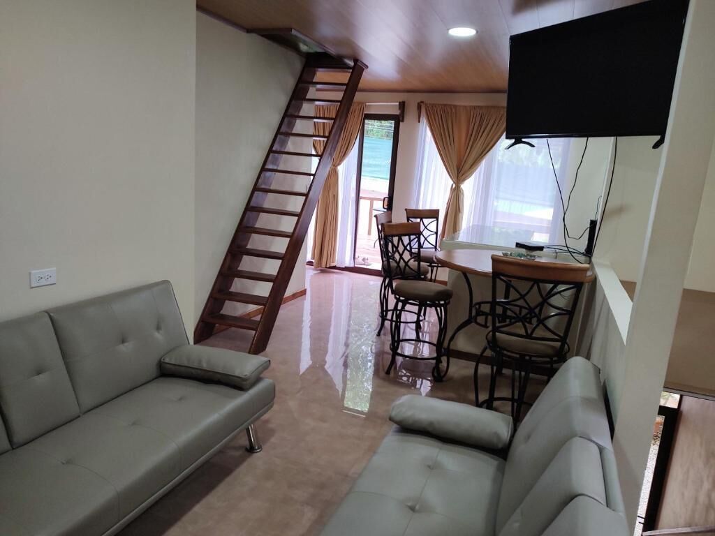 Un salon comprenant un canapé, une table à manger avec des chaises, un téléviseur fixé au mur et un escalier menant à un espace extérieur.