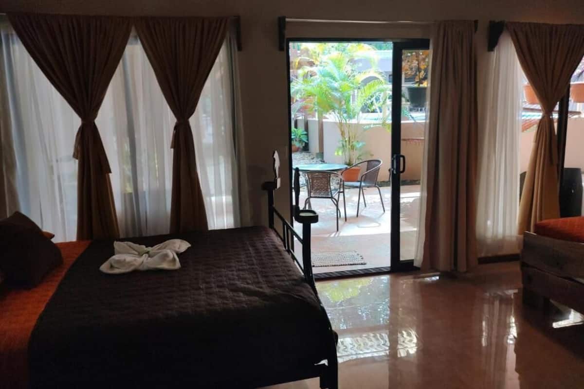 Une chambre d’hôtel avec un lit, une petite table et une chaise, près d’une porte-fenêtre coulissante donnant sur un espace extérieur agrémenté de plantes.
