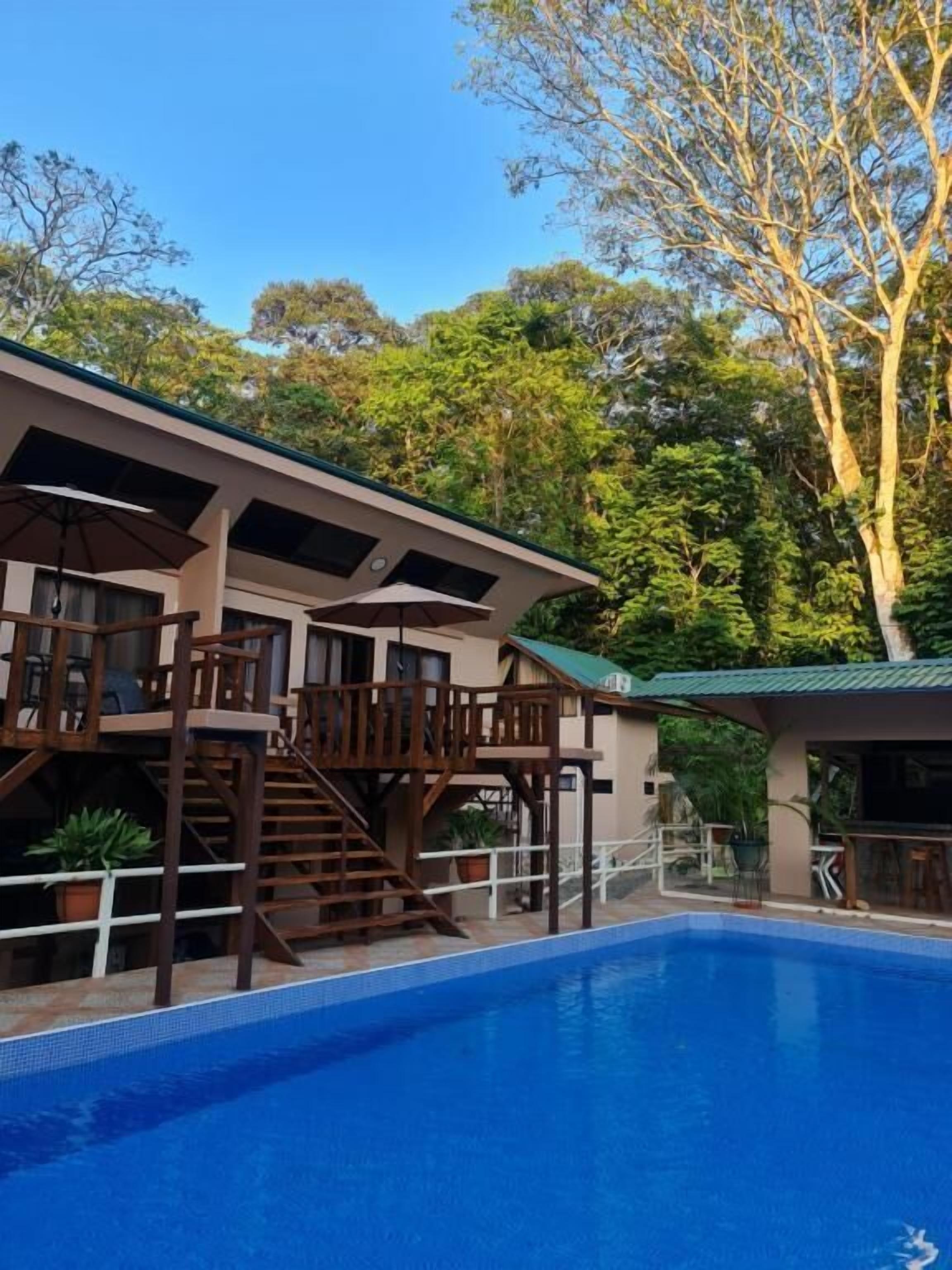 Un bâtiment à deux étages doté d’une piscine, d’une terrasse en bois et d’un espace extérieur aménagé pour s’asseoir.
