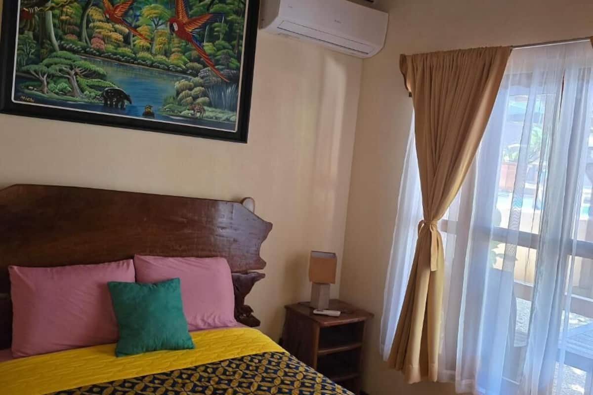 Une chambre à coucher avec une tête de lit en bois, un lit recouvert d’une literie à motifs, une table de chevet avec une lampe, un tableau représentant une scène de jungle et une fenêtre avec des rideaux.