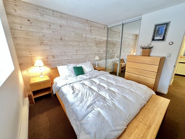 Un dormitorio con cabecera de madera, cama con ropa de cama blanca, mesita de noche con lámpara, cómoda de madera y un espejo en la pared.