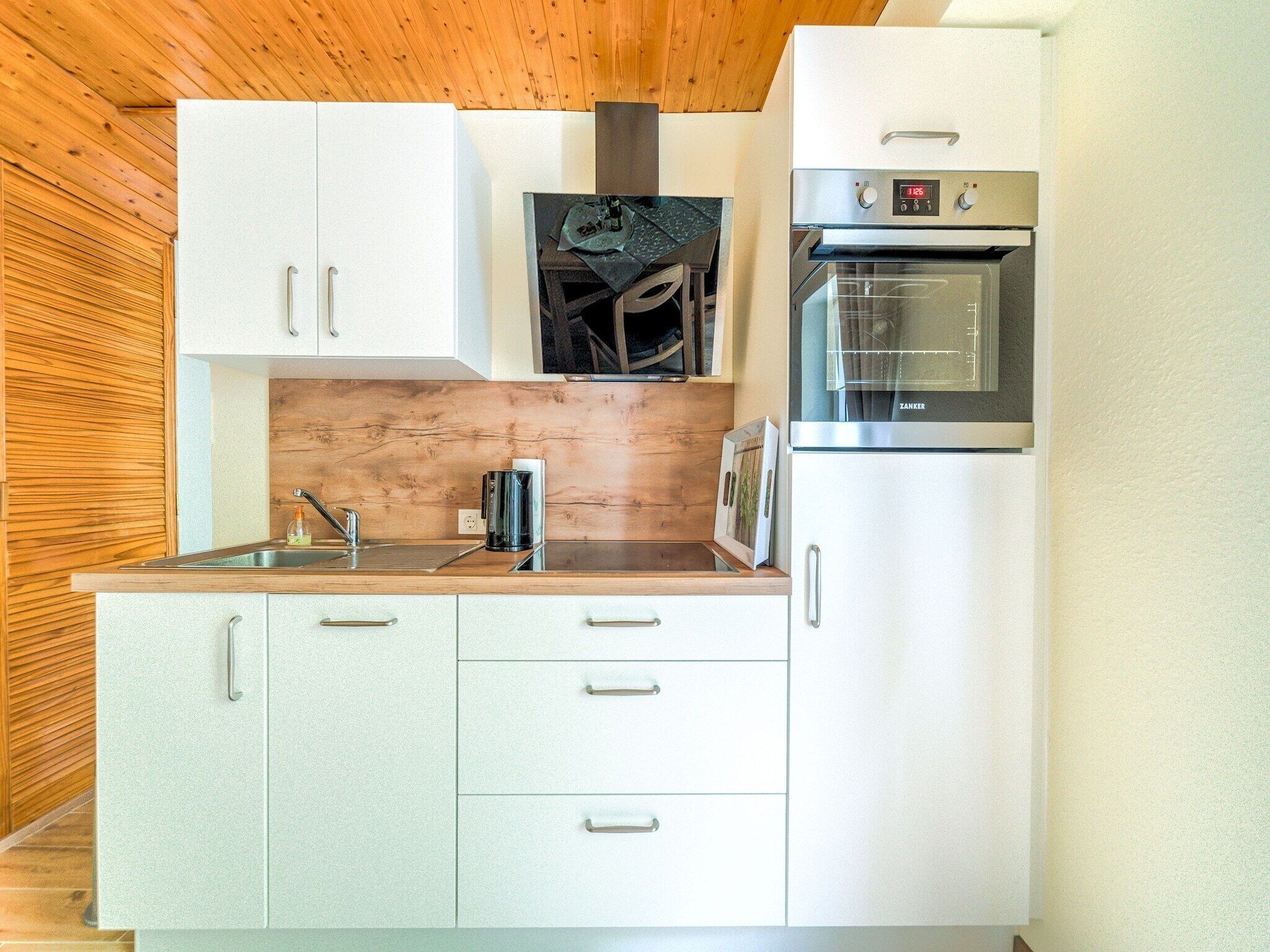 Cocina compacta con armarios blancos, horno de acero inoxidable y cafetera.