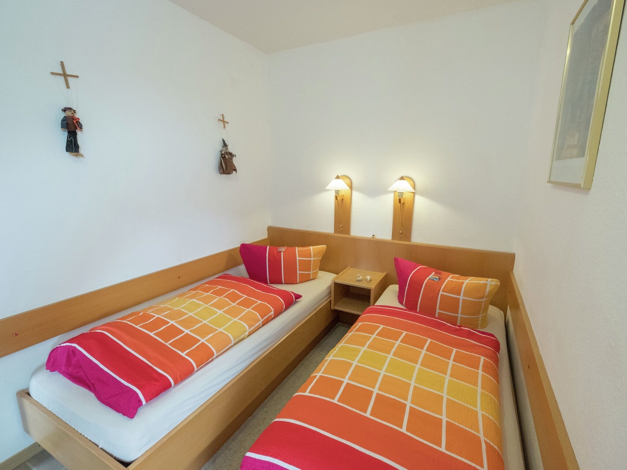 Een kamer met twee eenpersoonsbedden, elk met een houten hoofdbord en oranje beddengoed. Er staan twee nachtkastjes met lampen, en aan de muur hangt een kleine decoratie in de vorm van een kruis.