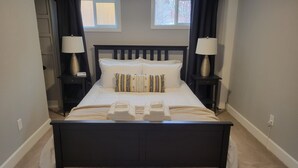 En pænt redt seng med hvide sengetæpper, to sengeborde med lamper og et vindue med gardiner.
