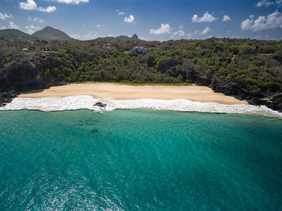 Un paysage côtier avec une plage de sable, des eaux turquoise et limpides, et une colline boisée en arrière-plan.