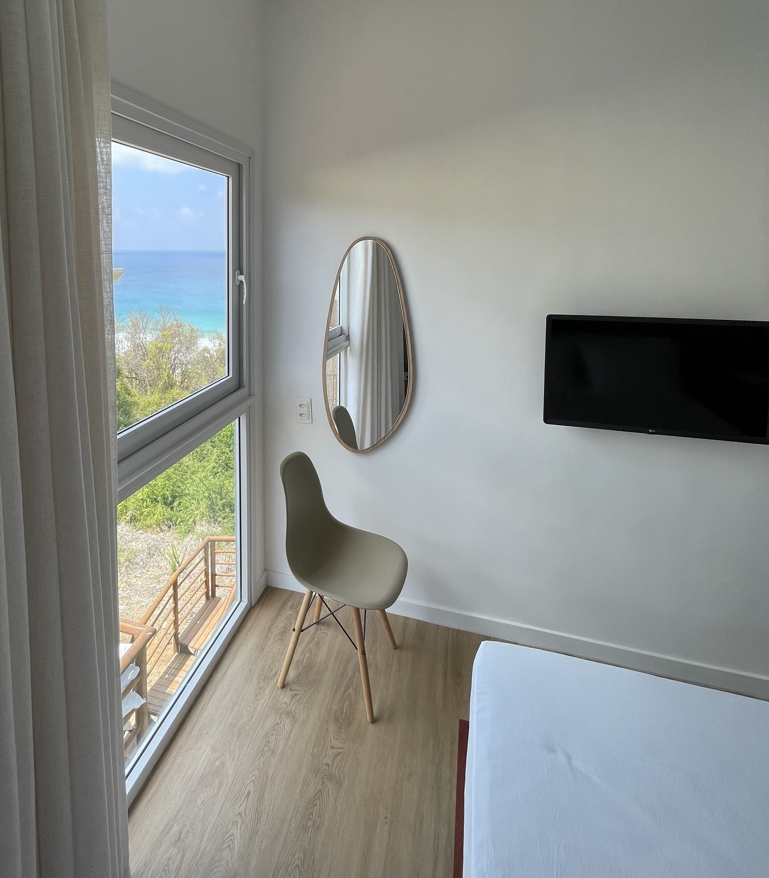 Un dormitorio con un ventanal grande, una silla, una cama, un televisor montado en la pared y un espejo.