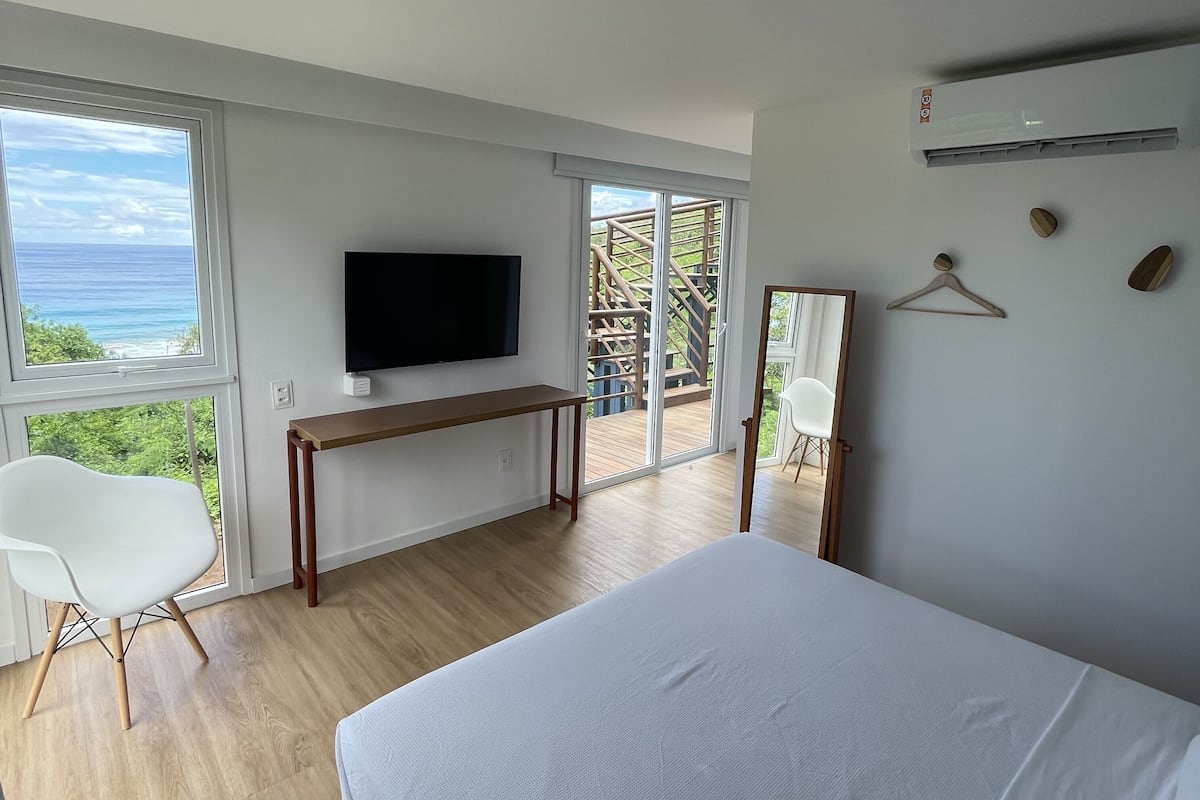 A bedroom with a large bed, a TV mounted on the wall, a wooden desk, and a balcony with a view of the ocean.