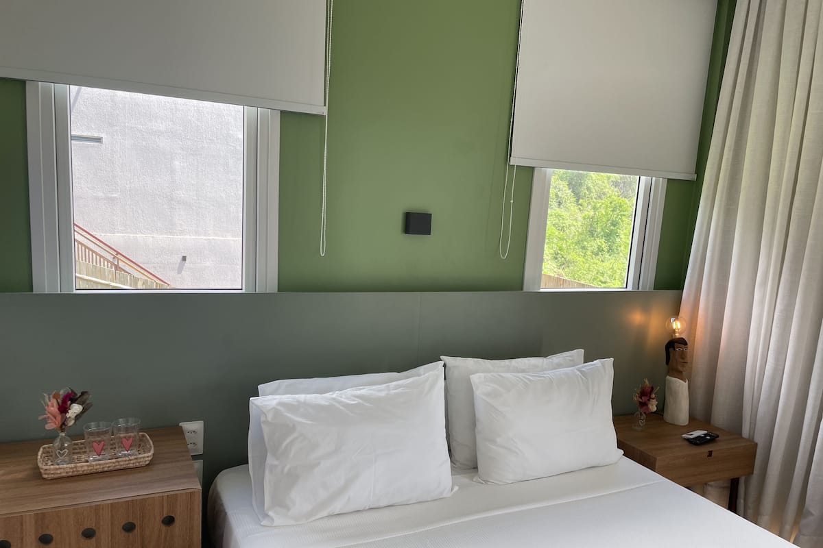 A bedroom with a bed, a nightstand, a window with blinds, and a view of a wooden staircase outside.