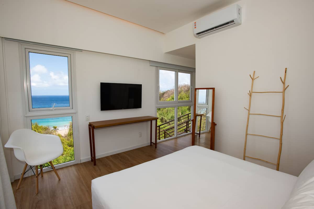 A bedroom with a large bed, a TV mounted on the wall, a desk, and a chair. There are large windows offering a view of the ocean and greenery.