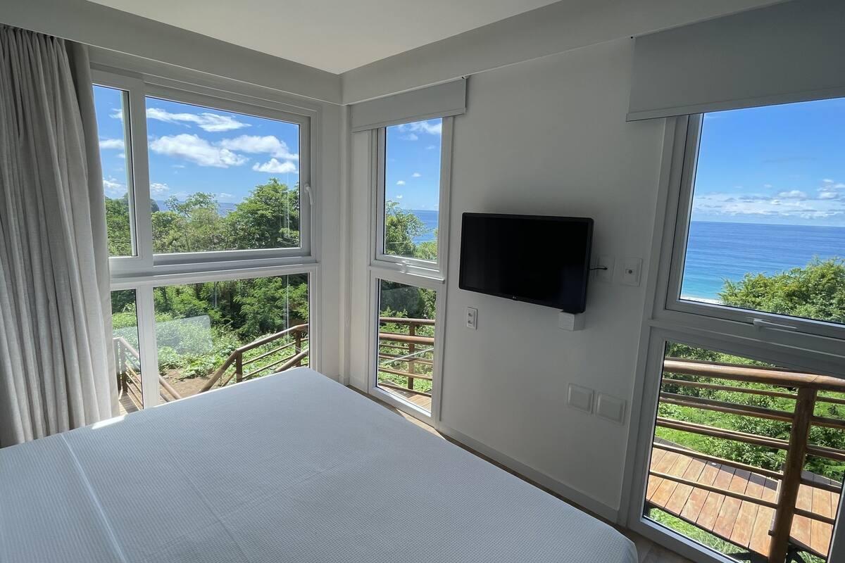 A bedroom with a large bed, a TV mounted on the wall, and large windows offering a view of the outdoors.