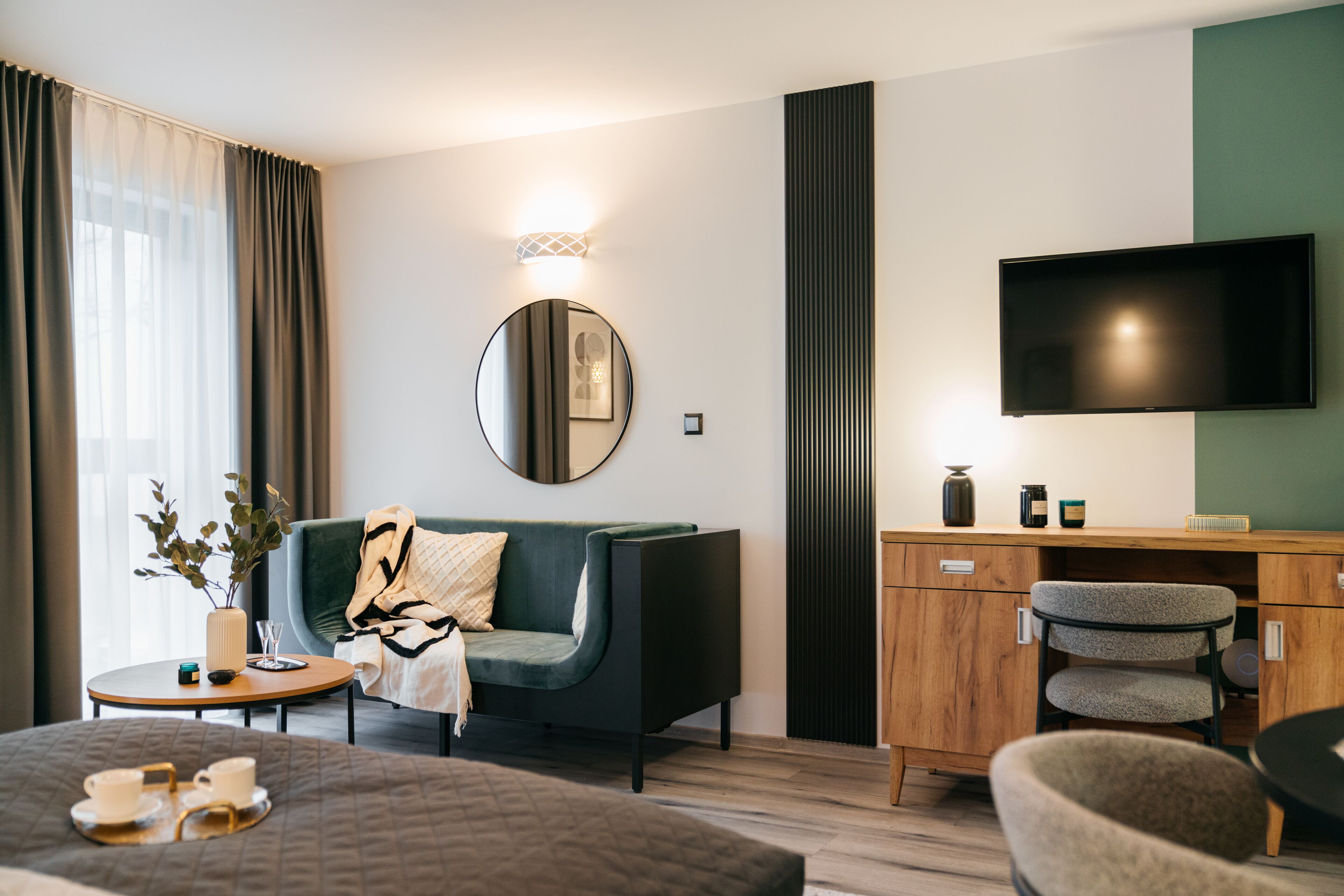 Una habitación de hotel moderna con una cama, un sofá, un televisor, un mueble de madera y un espejo redondo.