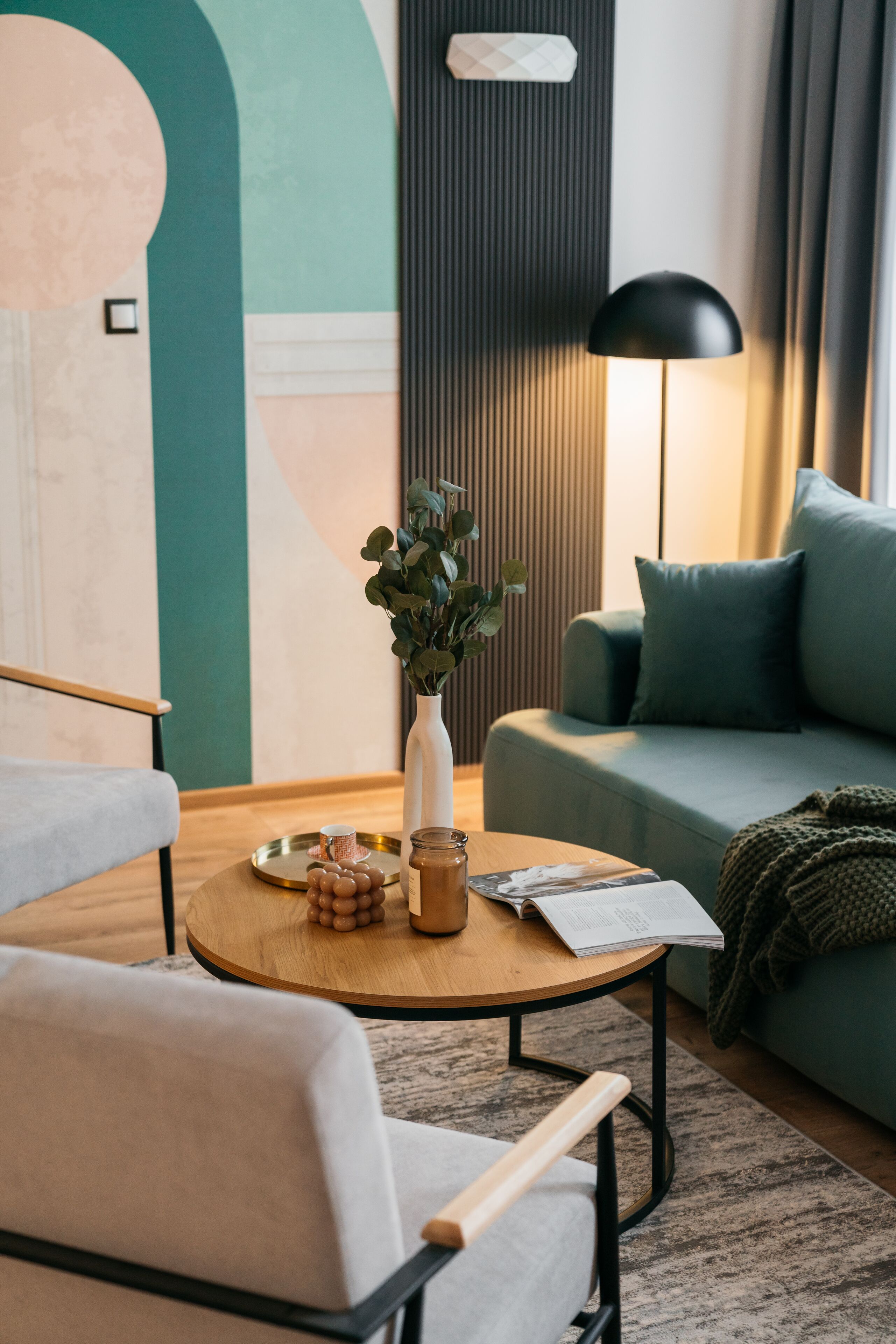 A modern living room with a round wooden coffee table, a white vase with green plants, a sofa with teal cushions, and a black floor lamp.