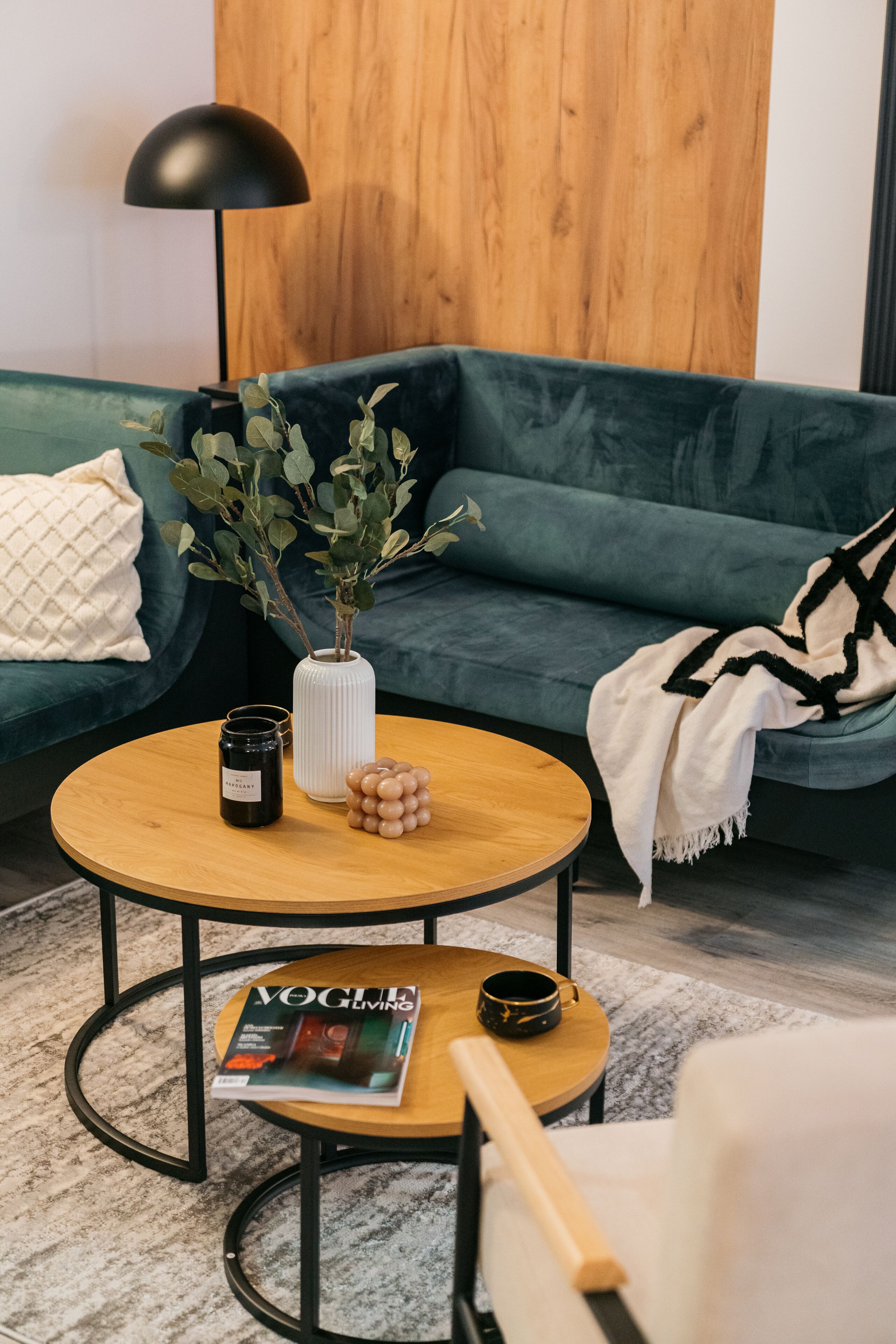 A modern living room with a green sofa, a round wooden coffee table, a white vase with green branches, a black candle, and a magazine on a small table.