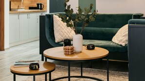 Una sala moderna con un sofá verde, una mesa de centro redonda y una mesita auxiliar. Un cuadro abstracto enmarcado adorna la pared, y se aprecia una cocina al fondo.