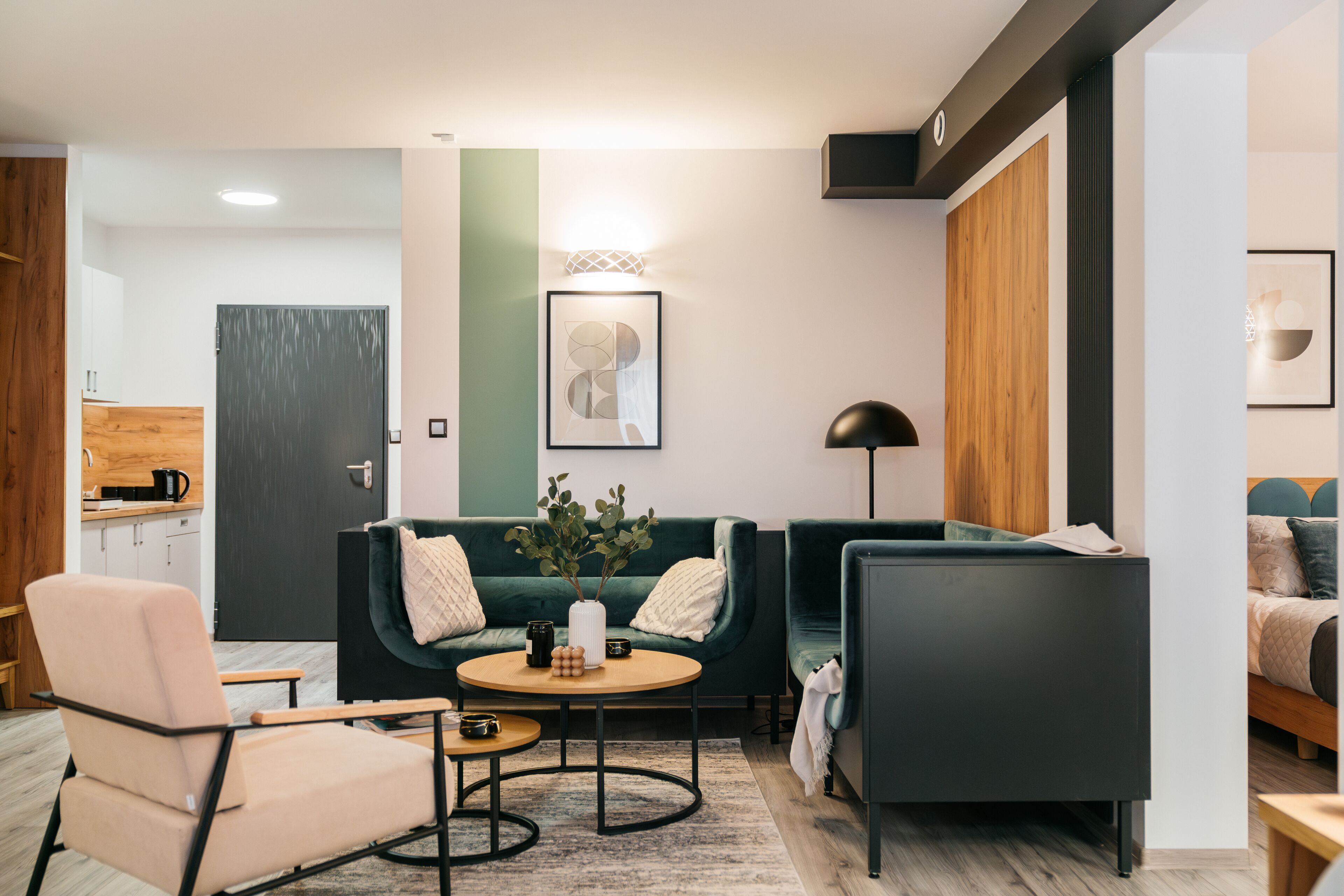 Ein modernes Wohnzimmer mit Sofa, Sessel und Couchtisch. Eine Tür führt in eine Küche, im Hintergrund ist ein Bett zu sehen.