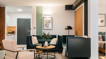 Ein modernes Wohnzimmer mit Sofa, Sessel und Couchtisch. Eine Tür führt in eine Küche, im Hintergrund ist ein Bett zu sehen.