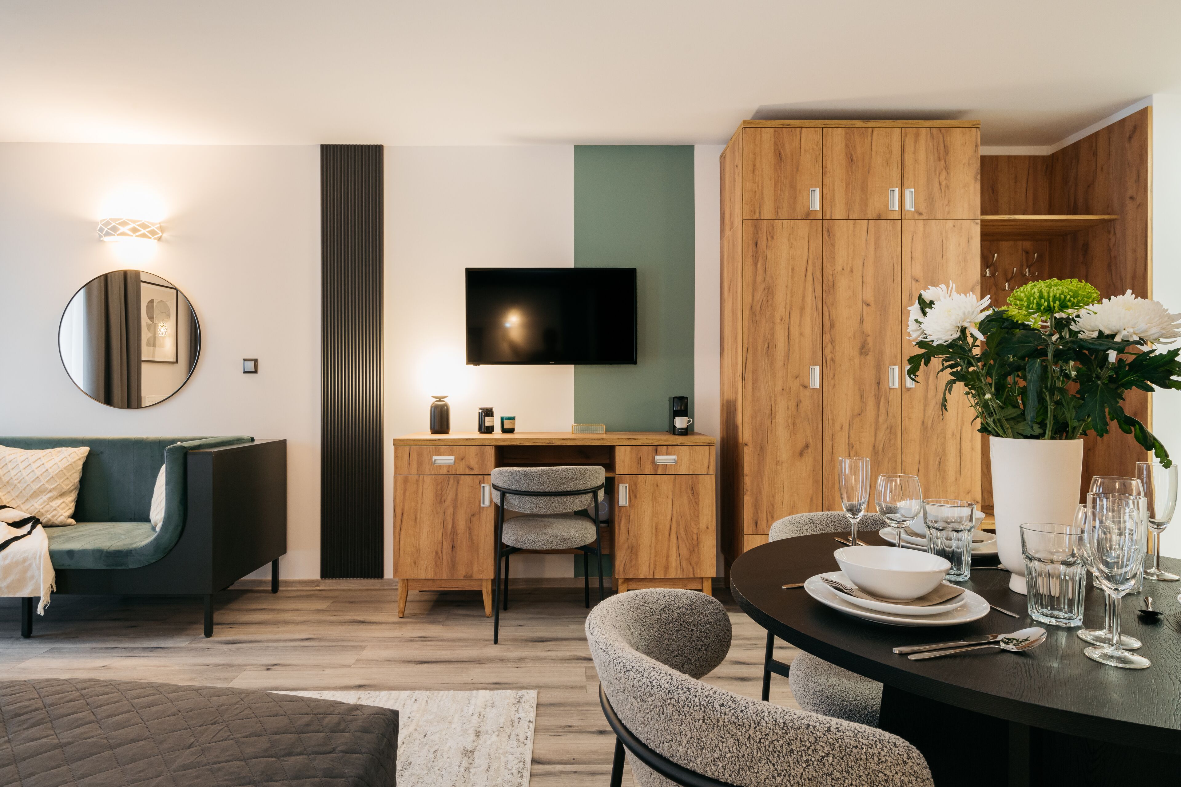 A modern living room with a sofa, a dining table set for four, a TV mounted on the wall, and wooden cabinets.