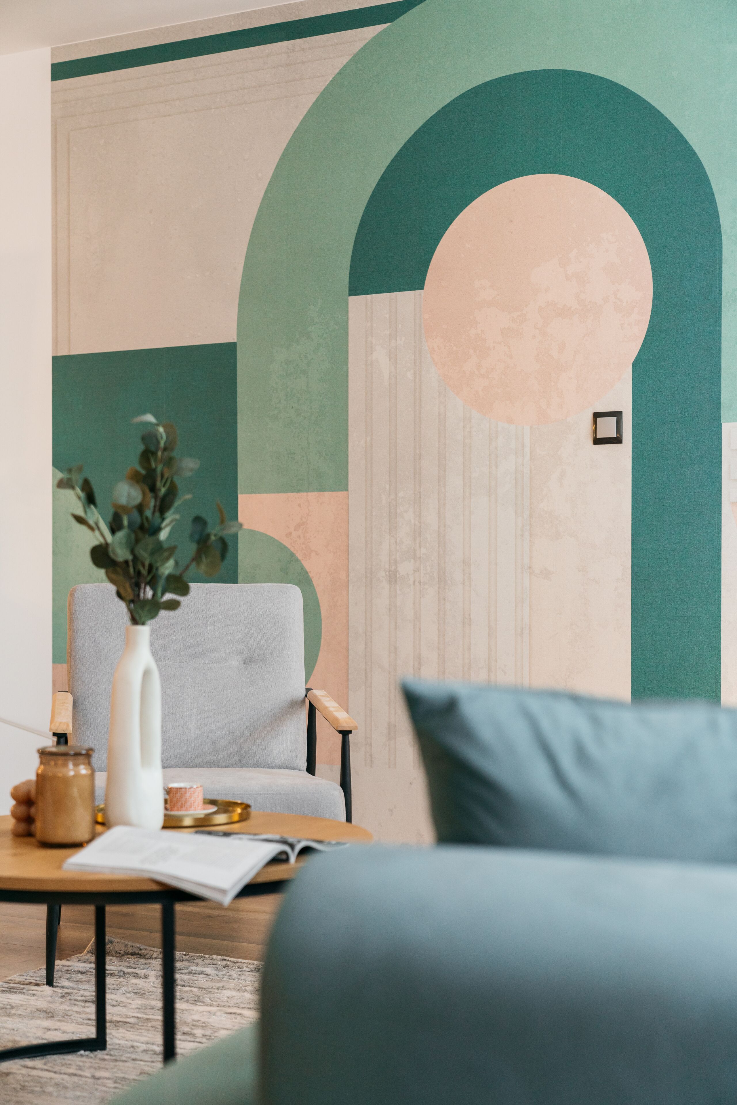 Una sala moderna con un sillón gris, una mesa de centro de madera con un florero de plantas verdes y un mural abstracto grande en la pared.