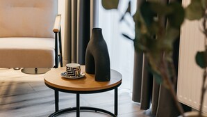Una sala moderna con un sofá, una mesa redonda de madera con un jarrón negro y una taza, y una lámpara de pie.