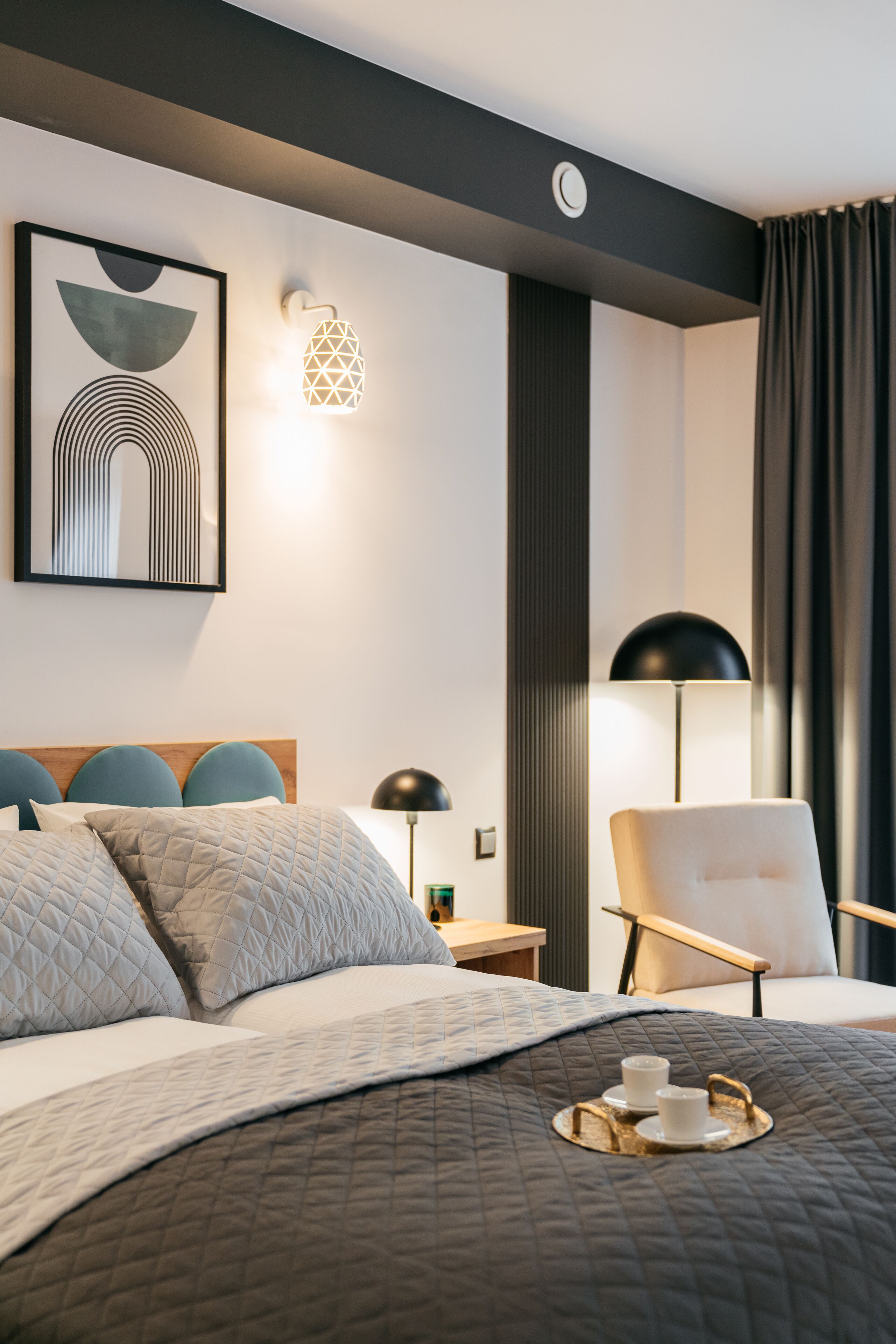 Ein modernes Hotelzimmer mit einem ordentlich bezogenen Bett, einem Nachttisch mit Lampe, einem Sessel und einer Wandleuchte.
