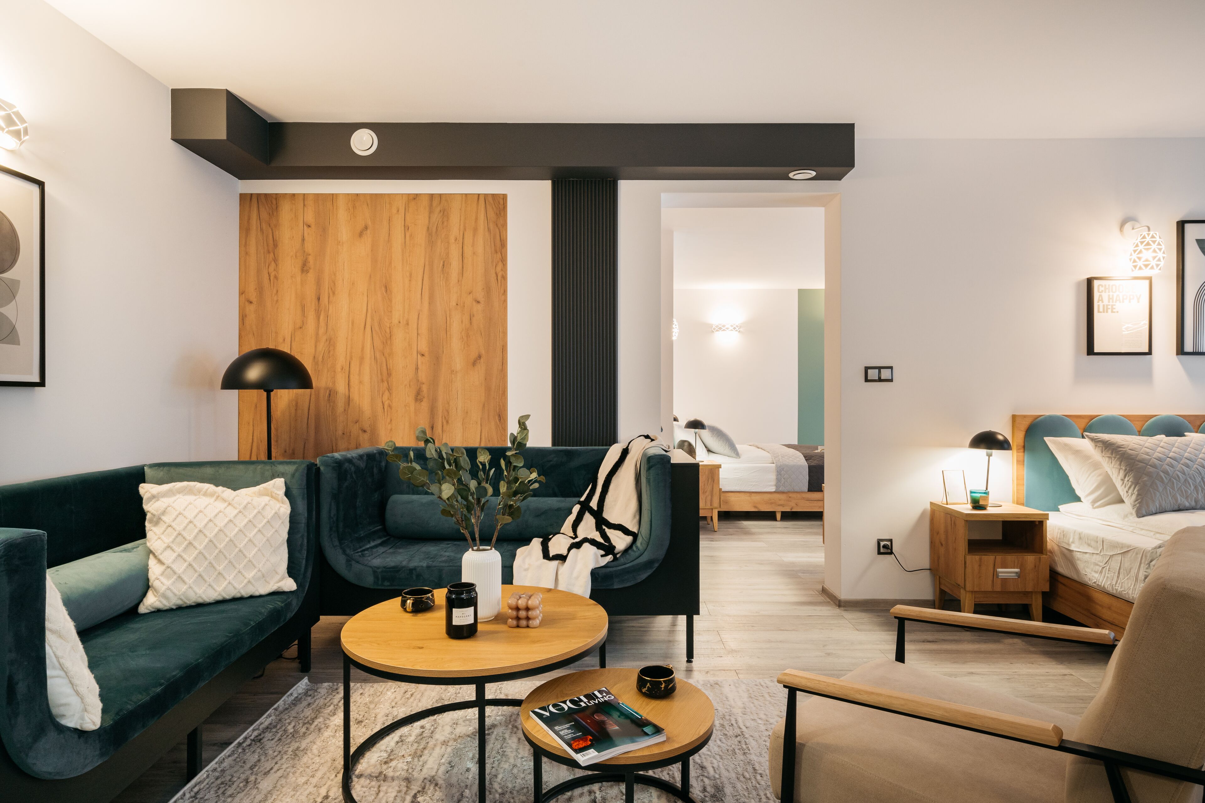Ein modernes Hotelzimmer mit einem grünen Sofa, einer Wand als Holzdekor, einem kleinen Holztischchen, einem Bett mit blauer Kopfteil und einem Nachttisch mit Lampe.