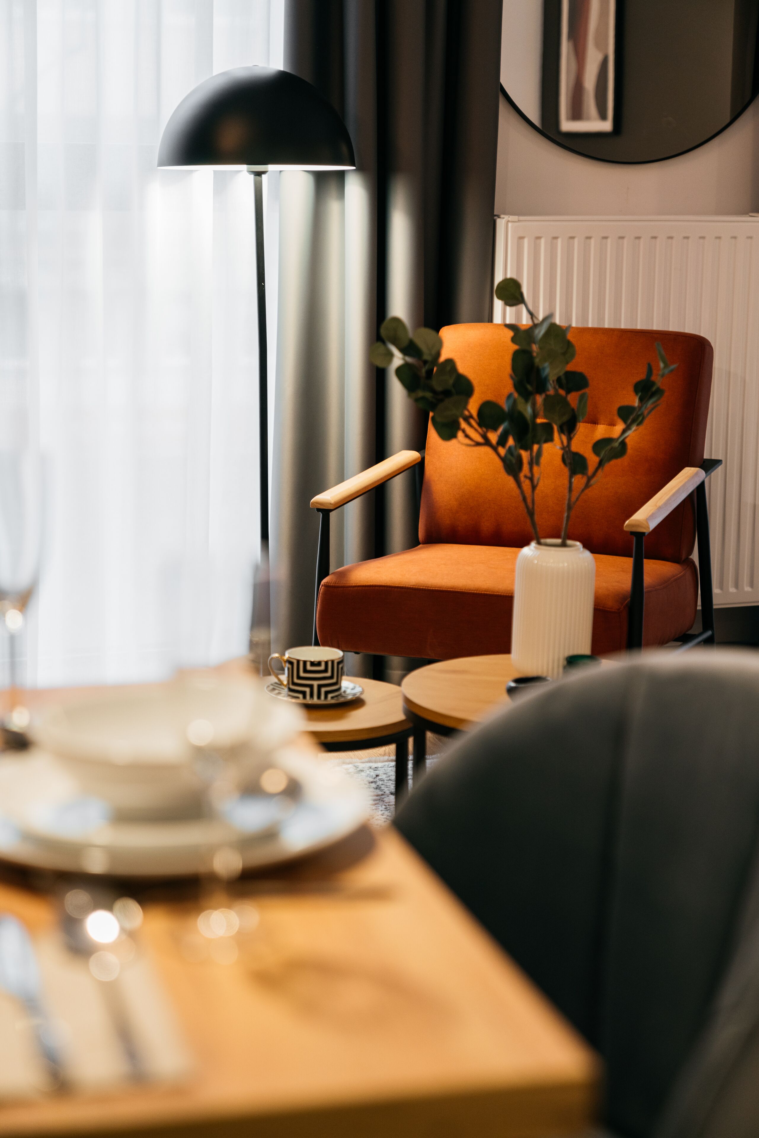 Ein modernes Zimmer mit einem Holztisch, einer schwarzen Stehlampe, einem orangefarbenen Sessel und einer weißen Vase mit grünen Blättern.