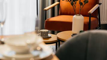 Ein modernes Zimmer mit einem Holztisch, einer schwarzen Stehlampe, einem orangefarbenen Sessel und einer weißen Vase mit grünen Blättern.