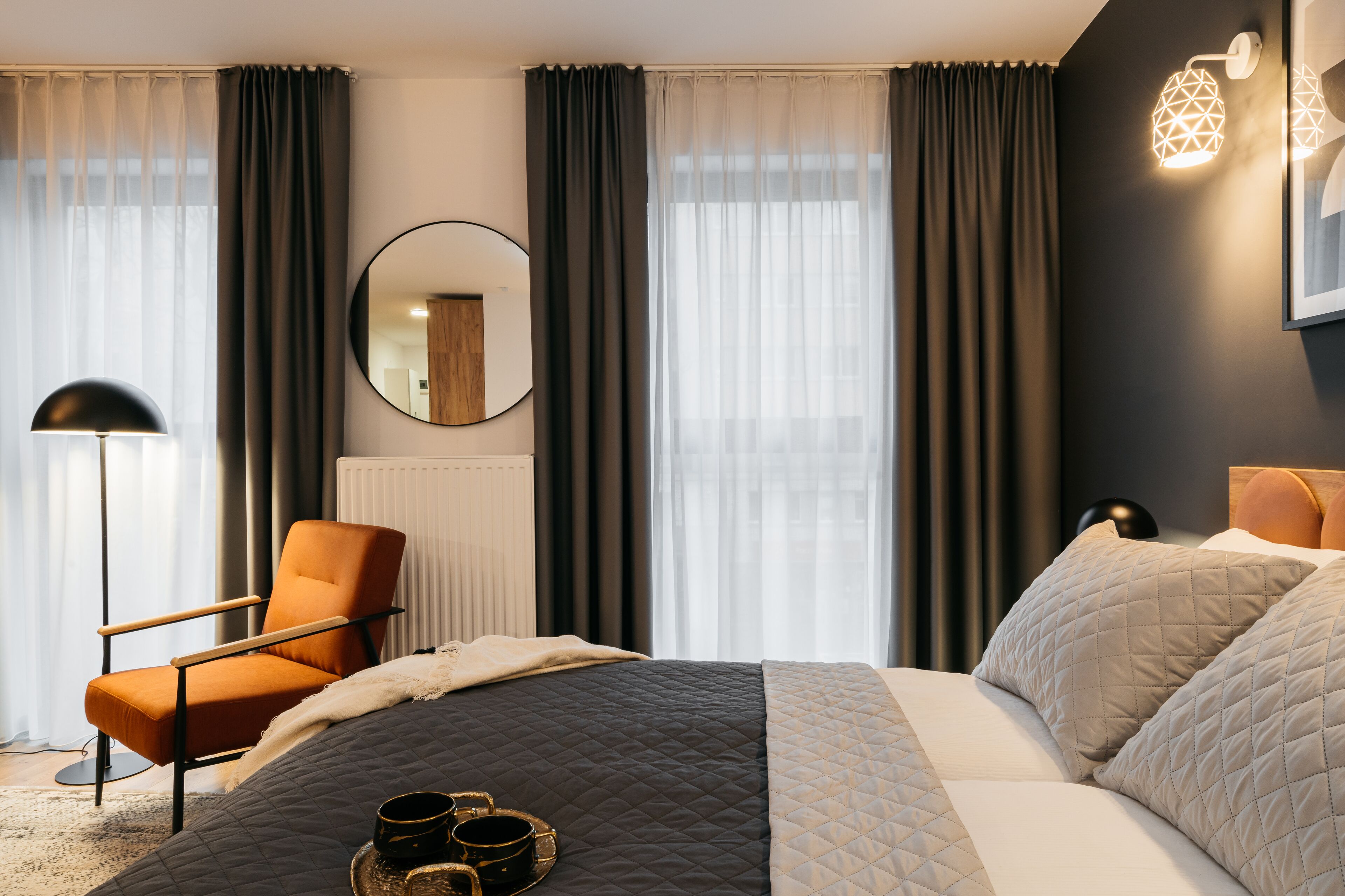 Une chambre d’hôtel moderne avec un grand lit, une chaise, un miroir rond et une vue sur la ville à travers des rideaux transparents.