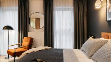 A modern hotel room with a large bed, a chair, a round mirror, and a view of the city through sheer curtains.