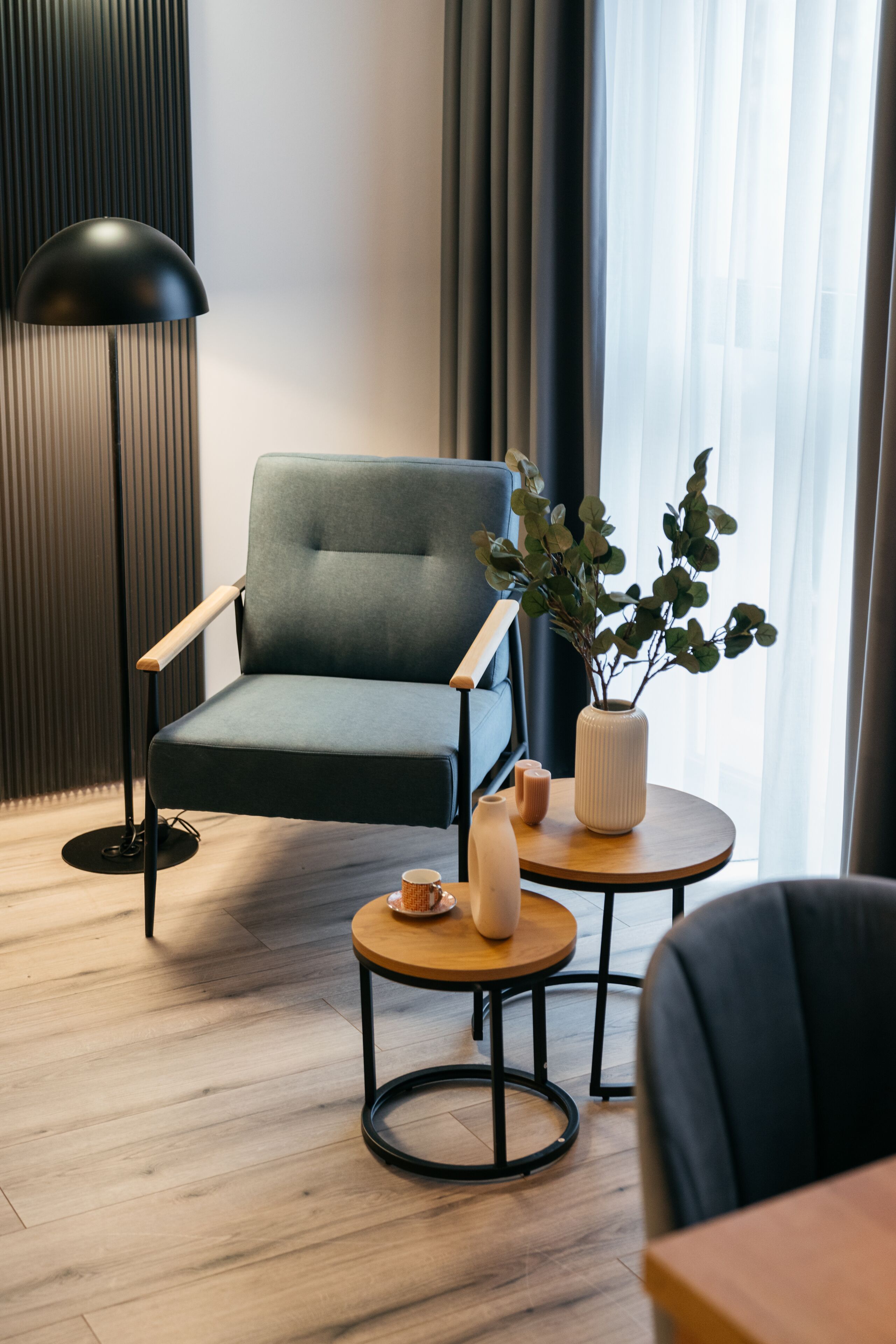 Una habitación moderna con un sillón azul oscuro, una lámpara de pie, una mesita redonda con un florero y una planta, y una mesa de centro de madera.