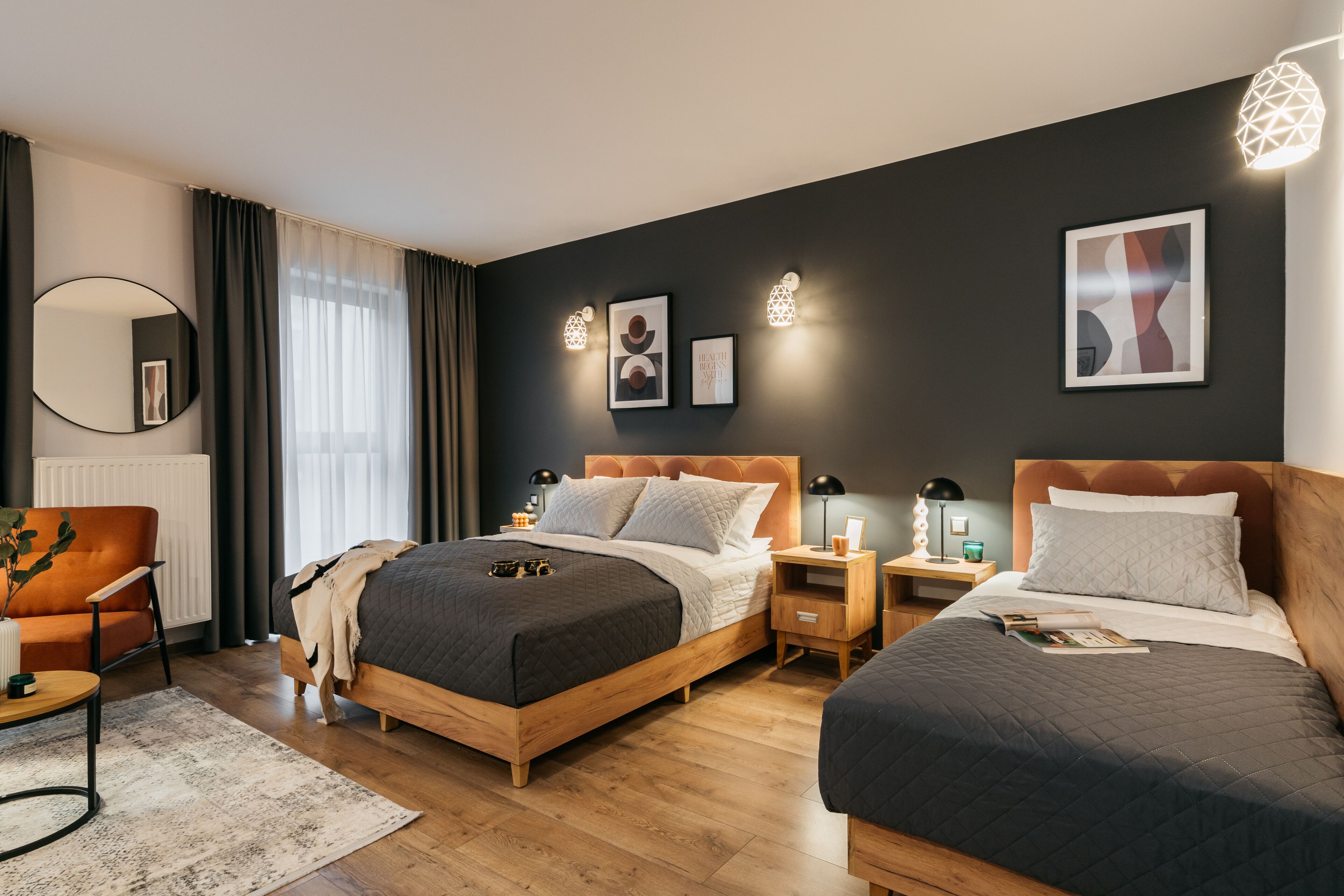 A modern hotel room with two beds, a round mirror, a wooden chair, and a bedside table with a lamp.