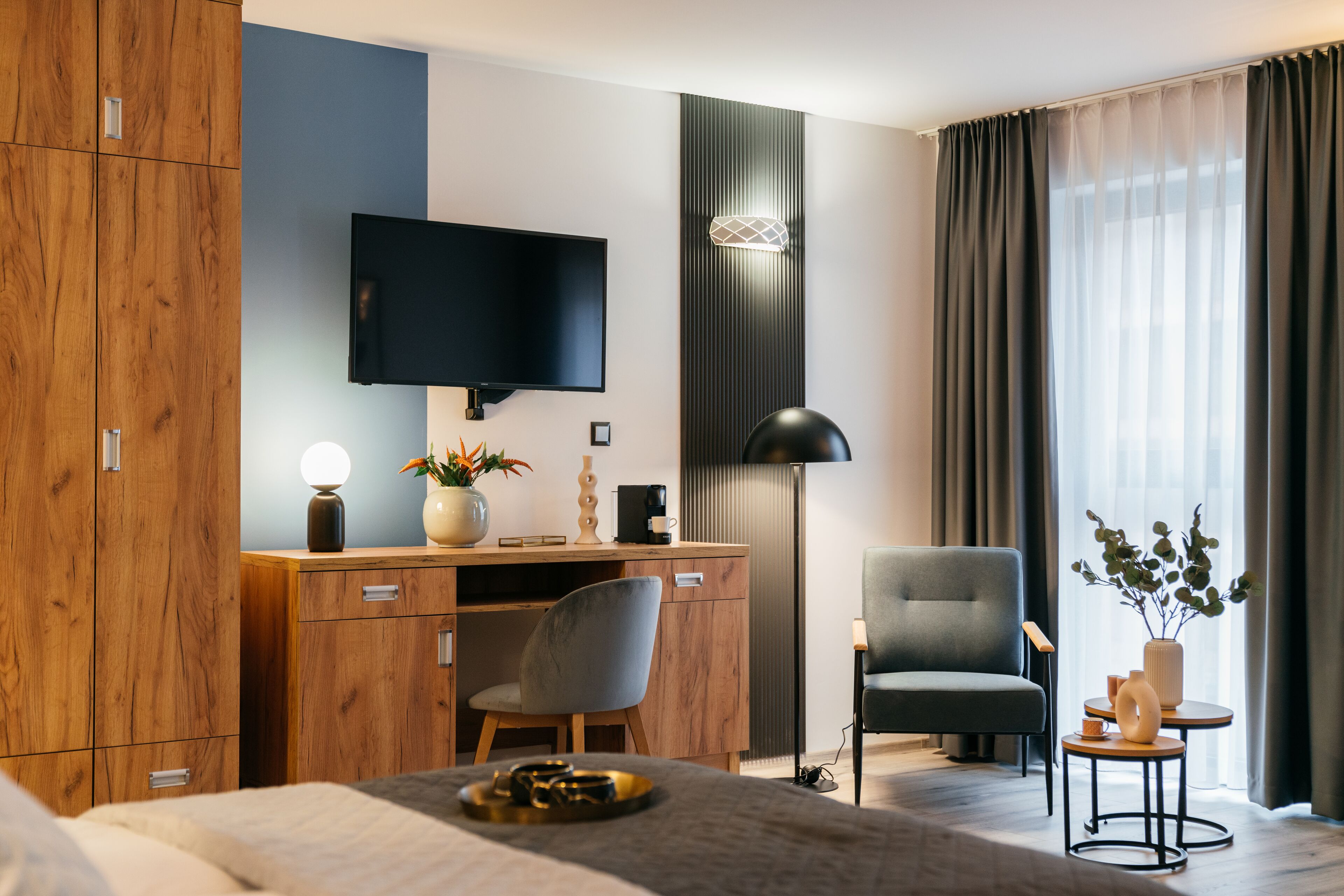 A modern hotel room with a wooden wardrobe, a desk with a flat-screen TV, a comfortable chair, a bed with a tray, and a small table with a vase.