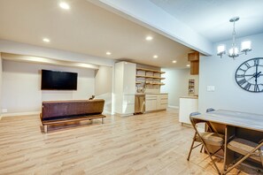 A modern living room with a flat-screen TV, a wooden bench, a dining table with chairs, and a kitchen area with cabinets and a clock on the wall.
