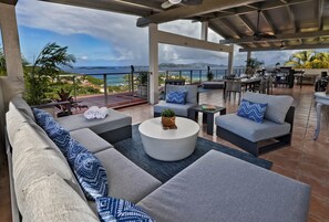A covered outdoor seating area with sofas, a round coffee table, and a view of the ocean.