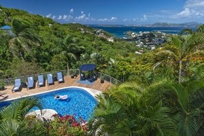 A swimming pool with lounge chairs, surrounded by lush greenery and overlooking a coastal view.
