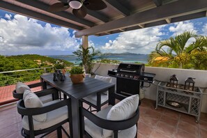 A patio with a table and chairs, a grill, and a view of the ocean and hills.