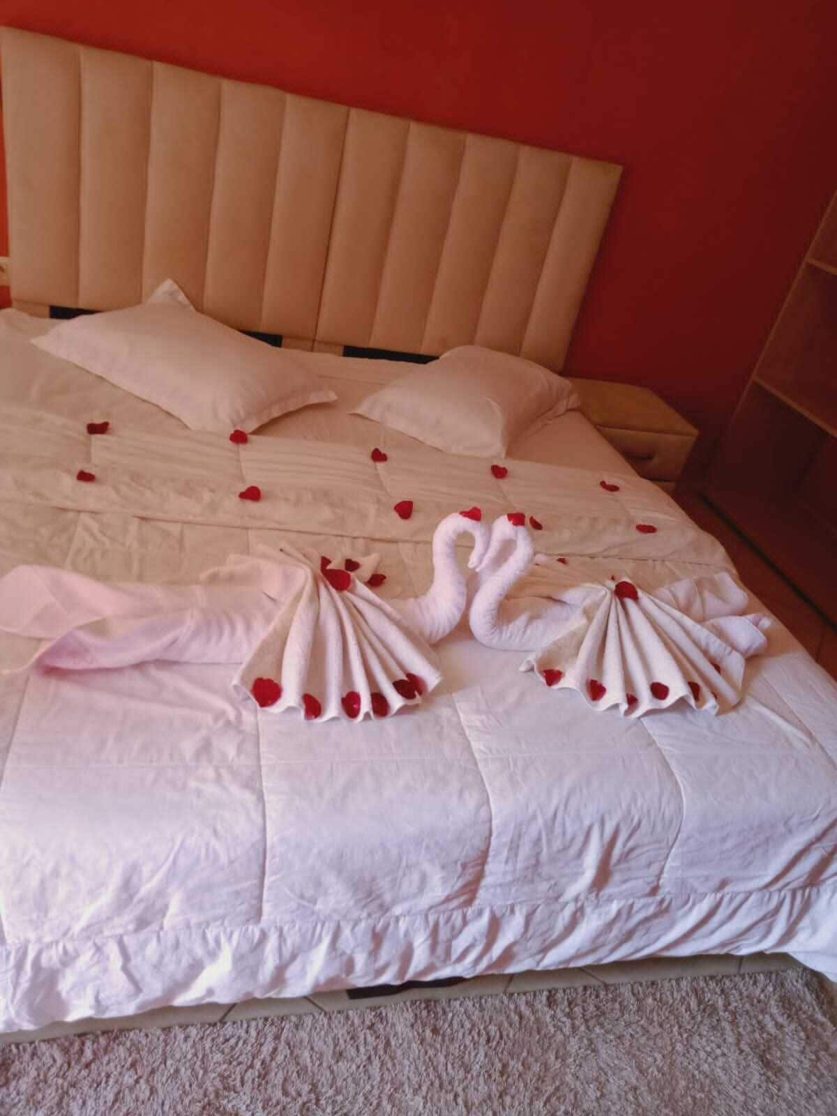 Una cama con sábanas blancas, pétalos de rosa roja y dos toallas con forma de cisne.