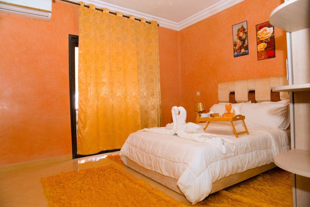 Un dormitorio con una cama, una mesita de noche de madera, una bandeja con objetos y una ventana con cortinas.