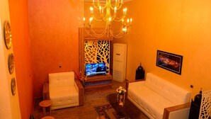 A living room with a white sofa, a white armchair, a wooden coffee table, a TV on a stand, and a chandelier. - Hotel citta rossa (Marrakech)
