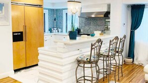 A modern kitchen with a white island, bar stools, a chandelier, and a large refrigerator.