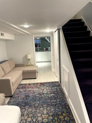 A living room with a sofa, a patterned rug, and a staircase leading to a kitchen area.