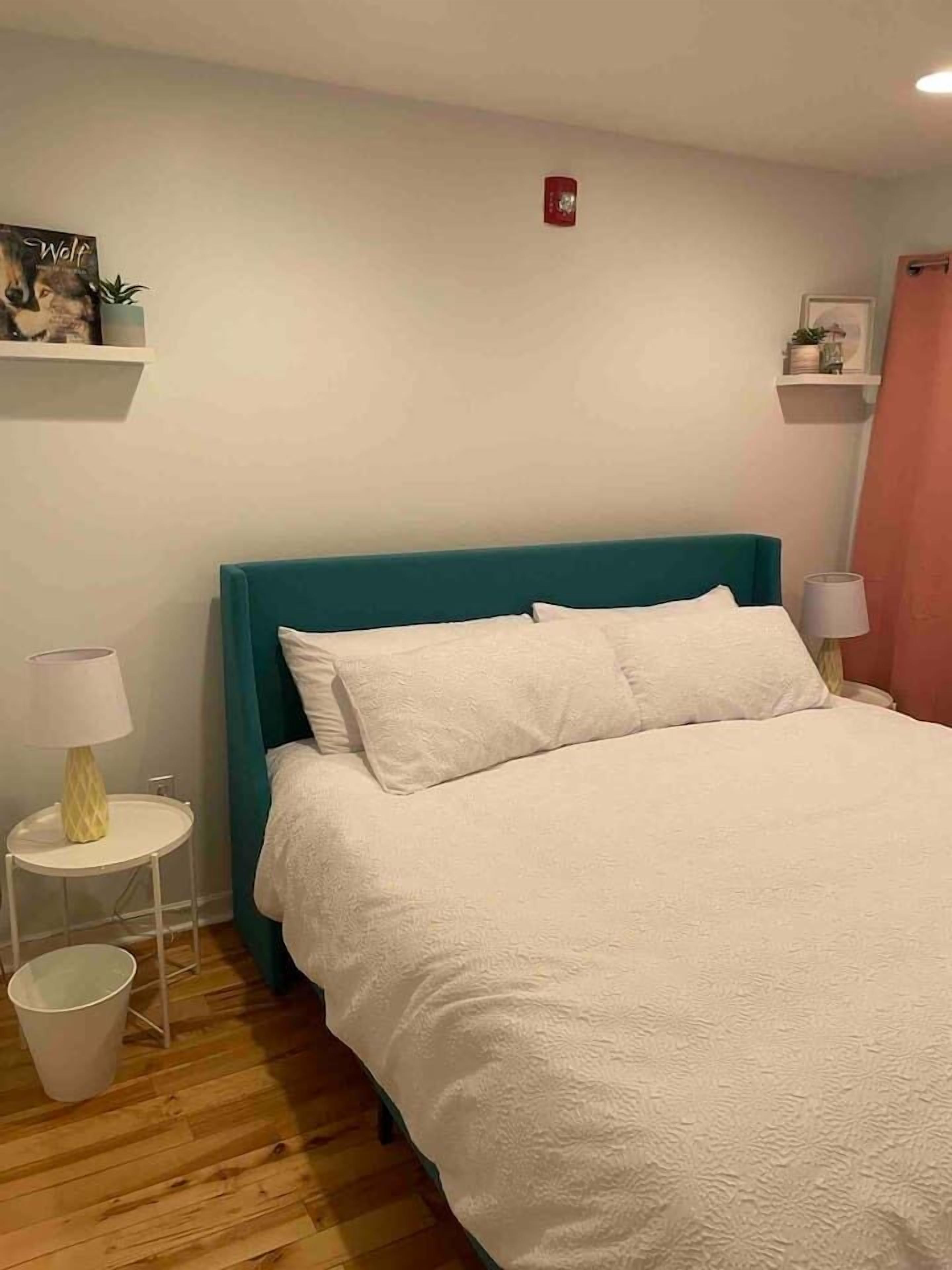 Una camera da letto con una testiera color petrolio, biancheria da letto bianca, un comodino con una lampada e una mensola a muro con una pianta.