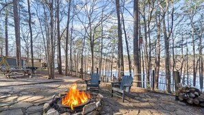 A stone fire pit surrounded by Adirondack chairs, a wooden deck, and a lake view. - Sunset Escape, Waterfront, Sleeps 6, Fishing, 2 Kayaks, Games, + Firepit! (Eatonton)